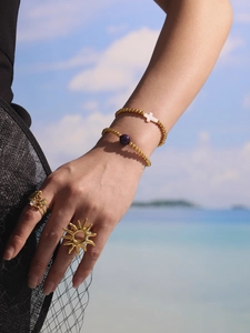 Detail of 18K gold-plated beaded bracelets with a cross charm and sun-shaped ring on beach