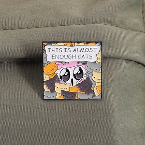 Detailed enamel pin with cartoon cats and text on olive green fabric background