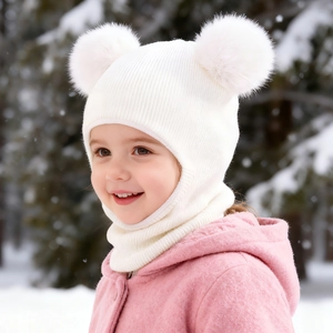 Detailed close-up of a white knit baby balaclava with dual fur pom-poms in snowy forest