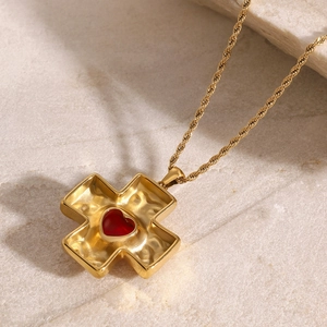 Detail shot of a gold cross pendant necklace featuring a red heart gemstone on marble
