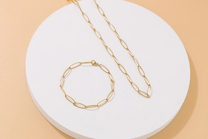 Golden paperclip chain necklace and bracelet set displayed on a white podium