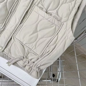 Detail of cream quilted puffer vest showing wavy stitching, patch pocket and adjustable drawstring
