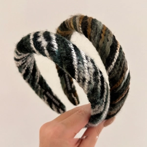 Hand holding two colorful braided knit headbands in striped wool against a beige background