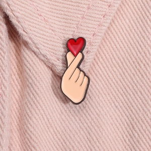 Close-up of a cartoon heart finger gesture enamel brooch on pink fabric background