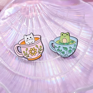 Pins mignons en émail avec chat et grenouille dans des tasses sur plateau violet