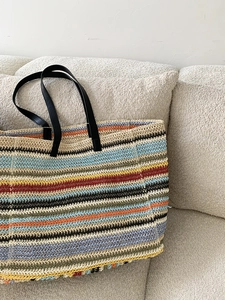 Large colorful striped straw tote bag with black handles on a minimalist sofa