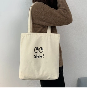 Detalle del bolso tote de lona blanco roto con texto shh negro y diseño de ojos animados