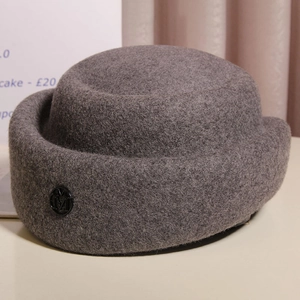 Close-up of dark grey wool felt pillbox hat with side metal logo button on a white table