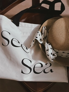 Detail of a beige canvas tote with See Sea print and a straw hat with a polka dot bow