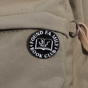 Close-up of black enamel Found Family Book Club pin badge on a khaki backpack strap