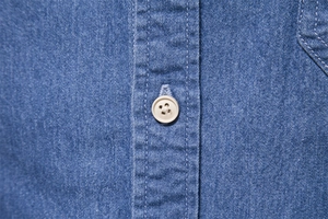 Macro detail of mid-blue denim shirt featuring white buttons and high-density stitching
