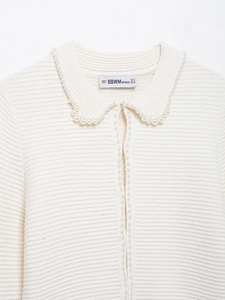 Détail du cardigan en maille blanche avec bordures en perles et étiquette BBWM