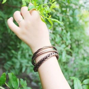 Dettaglio di bracciali intrecciati in bronzo e anelli lisci sul polso con sfondo verde natura