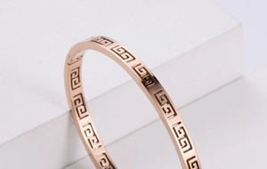 Close-up of rose gold hollow bangle featuring Greek key patterns on a geometric stand