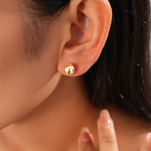 Close-up of a minimalist golden ball stud earring on a model's ear showing shiny texture