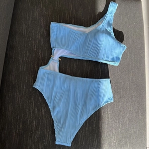 Detail of light blue ribbed one-shoulder swimsuit with side cutouts and gold metal rings