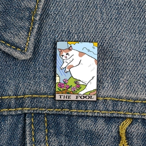 Macro detail of The Fool tarot cat enamel pin on dark denim jacket fabric