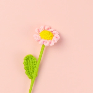 Macro view of a handmade crochet daisy with pink petals and green yarn stem on pink surface