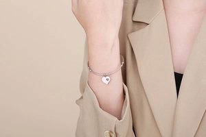 Detail shot of a silver heart charm bracelet on a model wearing a beige blazer