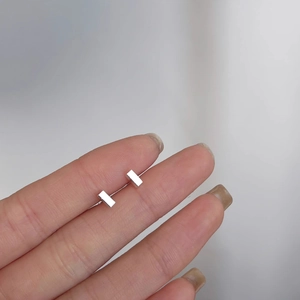 Close-up showing a pair of minimalist rectangular silver stud earrings held by hand