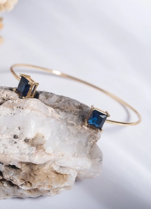 Detail showing gold open bangle featuring square deep blue gemstone inlays on white stone