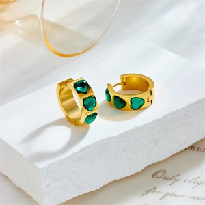 Golden heart-shaped hoop earrings with green emerald stones on a decorative white stone