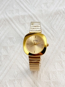 Detail of gold quartz watch with a square dial and flexible metal strap on fabric