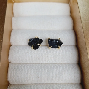 Natural black raw stone earrings with gold four-prong setting on beige textile background