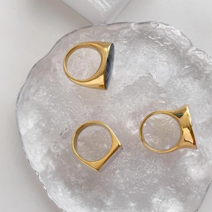 Detailed top and side views of chunky gold-plated signet rings on a textured glass tray