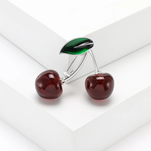 Elegant red cherry brooch with green enamel leaves on a white geometric platform