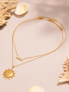 Detail van goudkleurige dubbele ketting met MOM- en zonhangers op een strandachtergrond