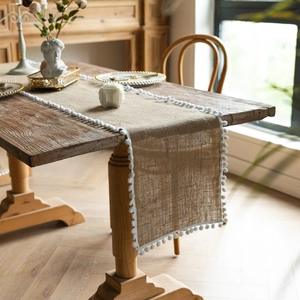 Close-up of a natural jute table runner with delicate white pom-pom lace trim on a wooden table