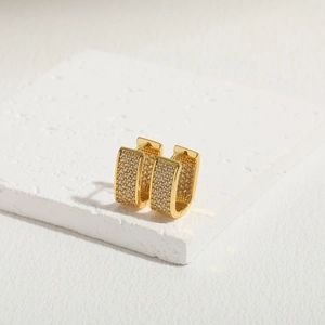 Macro detail of gold U-shaped earrings with square zircon pavé on white stone texture