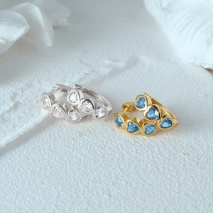 Detailed view of silver and gold heart huggie earrings with clear and blue zircon stones