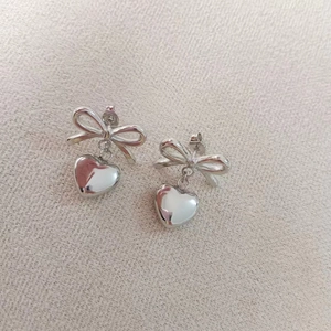 Close-up of silver bow stud earrings with heart pendants on textured fabric