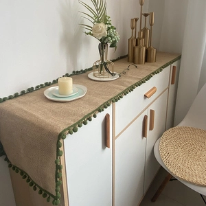 Natural jute table runner with dark green pom-pom trim displayed on a white cabinet