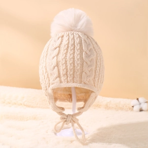 Detail of beige cable knit baby hat with ear flaps, white faux fur pompom and tie strings