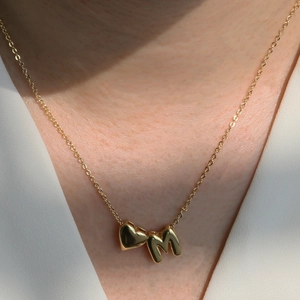 Close-up of golden 18k brass necklace featuring heart and letter M pendants worn by a model