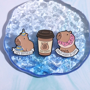 Three cute capybara enamel pins with food themes displayed on a blue water-textured tray