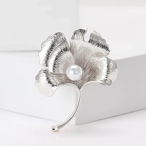 Macro detail of silver ginkgo leaf brooch with central pearl inlay on white background
