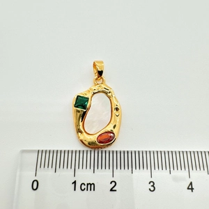 Macro shot of gold-plated oval pendant with green and red gemstones next to a ruler