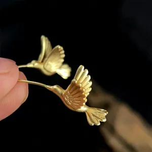Artistic hummingbird stud earrings in matte gold finish held in hand against dark backdrop