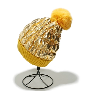 Detailed shot of golden coated knit hat with yellow faux fur pompom and ribbed brim