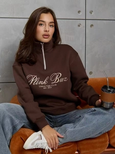 Détail du sweat marron foncé à quart de zip avec logo blanc porté sur un canapé orange