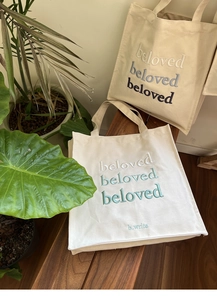 Detail of embroidered beloved text on large canvas tote bags on a wooden bench