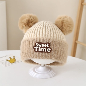 Dettaglio del berretto in maglia beige con due pompon e una toppa con lettere Sweet Time