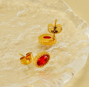 Dettaglio macro di orecchini ovali dorati con zircone rosso, bordo milgrain e chiusura a farfalla