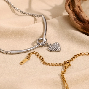 Détail du pendentif cœur argenté avec strass et chaîne Figaro dorée sur tissu beige doux