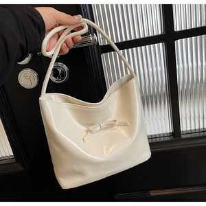 Creamy white patent leather bucket bag with a bow accent held by a person in a stylish setting