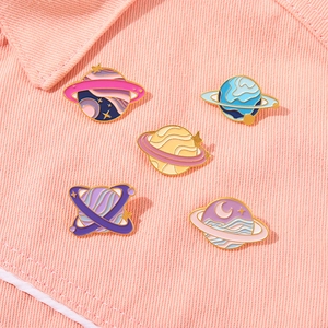 Five assorted colorful planet enamel brooches pinned on pink denim fabric material